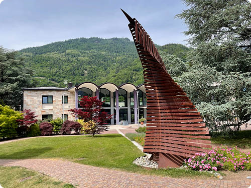 Vor einem modernen Geb ude mit gro en Fenstern steht eine abstrakte, rostbraune Metallskulptur in Form eines aufsteigenden, gezackten Segels, umgeben von gr nen B umen und Blumen. Beschreibung von KI generiert