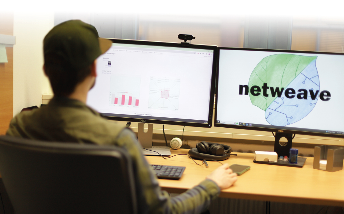 Eine Person sitzt an einem Schreibtisch vor zwei Monitoren, auf einem ist das Wort „netweave“ mit einem Blattsymbol zu sehen, w hrend der andere Diagramme anzeigt. Beschreibung von KI generiert