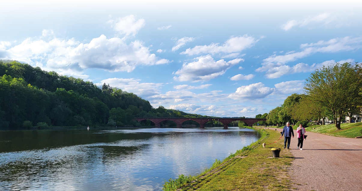 Uferpromenade an einem breiten Fluss bei klarem Wetter. Rechts verl uft ein breiter, kiesiger Fu weg, auf dem zwei Personen nebeneinander spazieren; eine f hrt an einer kurzen Leine einen kleinen Hund. Links breitet sich ruhiges Wasser mit leichter Spiegelung des Himmels aus. In der Bildmitte im Hintergrund berspannt eine rotbraune Steinbogenbr cke den Fluss. Linkes Ufer und die Flussufer im Hintergrund sind dicht mit gr nen B umen und bewaldeten H geln bedeckt. Der Himmel ist blau mit vielen wei en, leicht verstreuten Wolken. Stimmung: entspannter, sonniger Sp tnachmittag an der Flusslandschaft.