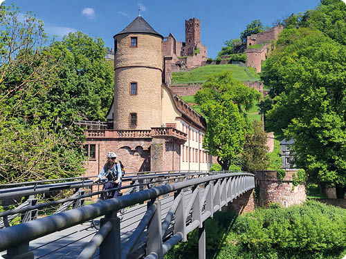 Blick ber eine schmale Fu - und Radbr cke auf ein historisches Burggel nde. Im Vordergrund kommt von links eine Person auf dem Fahrrad auf die Kamera zu; das Gesicht ist unscharf/abgeblendet, die Person tr gt Helm, Rucksack und freizeittaugliche Kleidung. Die Br cke hat graue Gel nder und f hrt zu einem l nglichen, hellen Geb ude mit einem runden, beigen Turm an der linken Seite. Hinter dem Geb ude steigen bewachsene Hangterrassen und Burgmauern aus r tlichem Stein auf; weiter oben sind Ruinenmauern und ein quadratischer Turm sichtbar. Rechts und links der Szene dichte, gr ne B ume; der Himmel ist blau mit wenigen Wolken. Stimmung: sonniger Fr hlingstag, touristisch-historische Atmosph re.