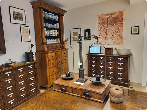 Innenaufnahme eines historischen Apotheken- oder Kr merladens. Im Vordergrund auf einem Holztisch steht eine alte Balkenwaage mit zwei runden Schalen; die Waage ist auf einer flachen Holztruhe mit zwei schwarzen Griffen befestigt. Rechts auf dem Tisch liegen zwei feste Kn uel Bindfaden und zwei messingfarbene Gewichtszylinder. Im mittleren Bildbereich zwei niedrige Schubladenschr nke nebeneinander, jeder mit vielen kleinen Holzschubladen und wei en Beschriftungsstreifen. Dazwischen ein hoher Holzschrank mit vielen Schubladen und oben ein Aufsatz-Regal, in dem blau-wei e Keramikgef e stehen. An der rechten Wand ein gr  eres Flachrelief oder Druck mit historischem Stadtbild. An der linken Wand gerahmte Dokumente und Notizen. Die Atmosph re wirkt ruhig, gut erhalten und museal; Materialien haupts chlich dunkles Holz und Keramik.