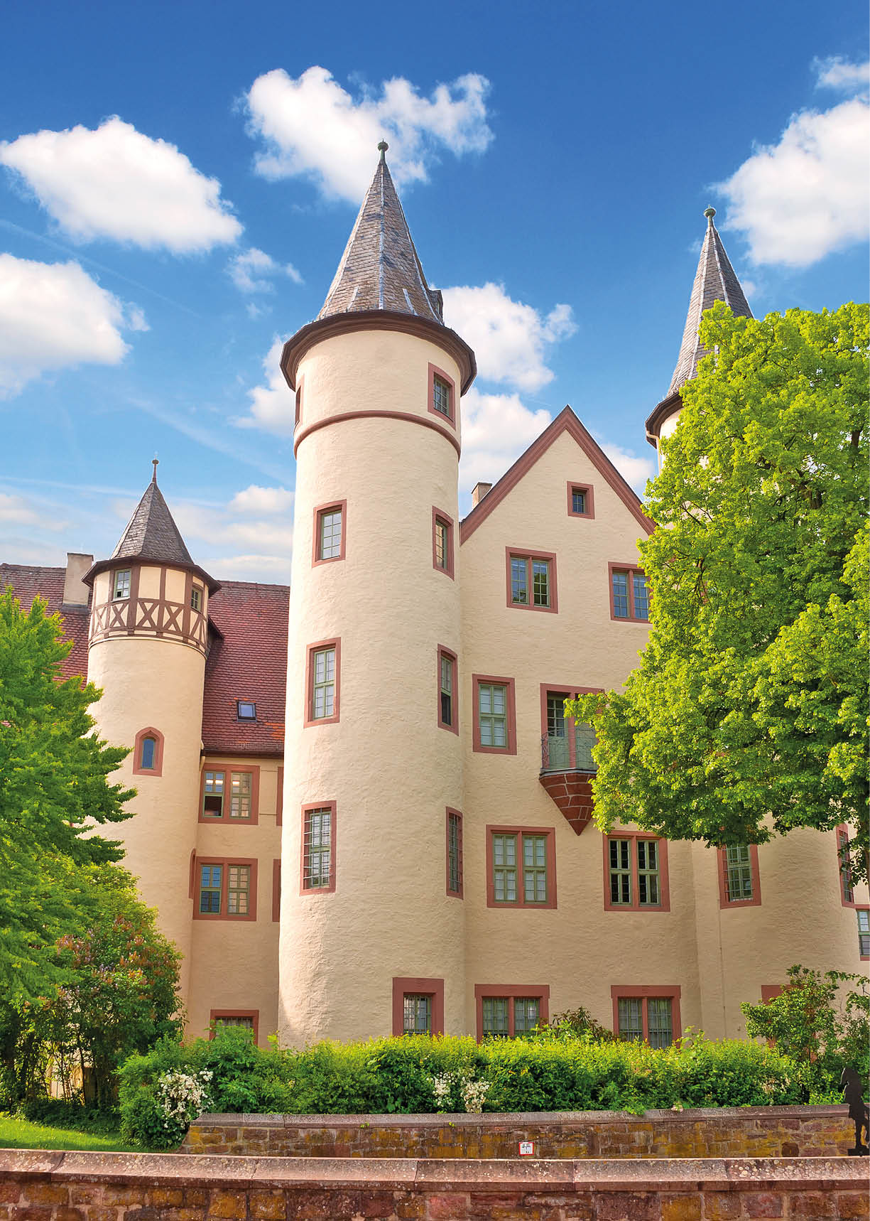Aufnahme einer hellen, gut erhaltenen Schlossfassade mit mehreren runden T rmen und spitzen D chern. Im Vordergrund eine niedrige, rote Bruchsteinmauer und ein Streifen gepflegter Bepflanzung mit Str uchern. Dahinter erhebt sich das cremefarbene Geb ude: drei prominente runde T rme mit schiefergedeckten Kegeld chern, zwischen ihnen ein dreist ckiger Mittelbau mit geneigtem Dach und rechteckigen Fenstern. Der Turm links zeigt zus tzlich Fachwerk oberhalb des Rundbaus. Rechts wachsen hohe, gr ne Laubb ume bis in die Fassadenh he. Der Himmel ist klarblau mit einigen wei en Wolken. Stimmung: sonniger Fr hlingstag, architektonisch ruhig und freundlich.