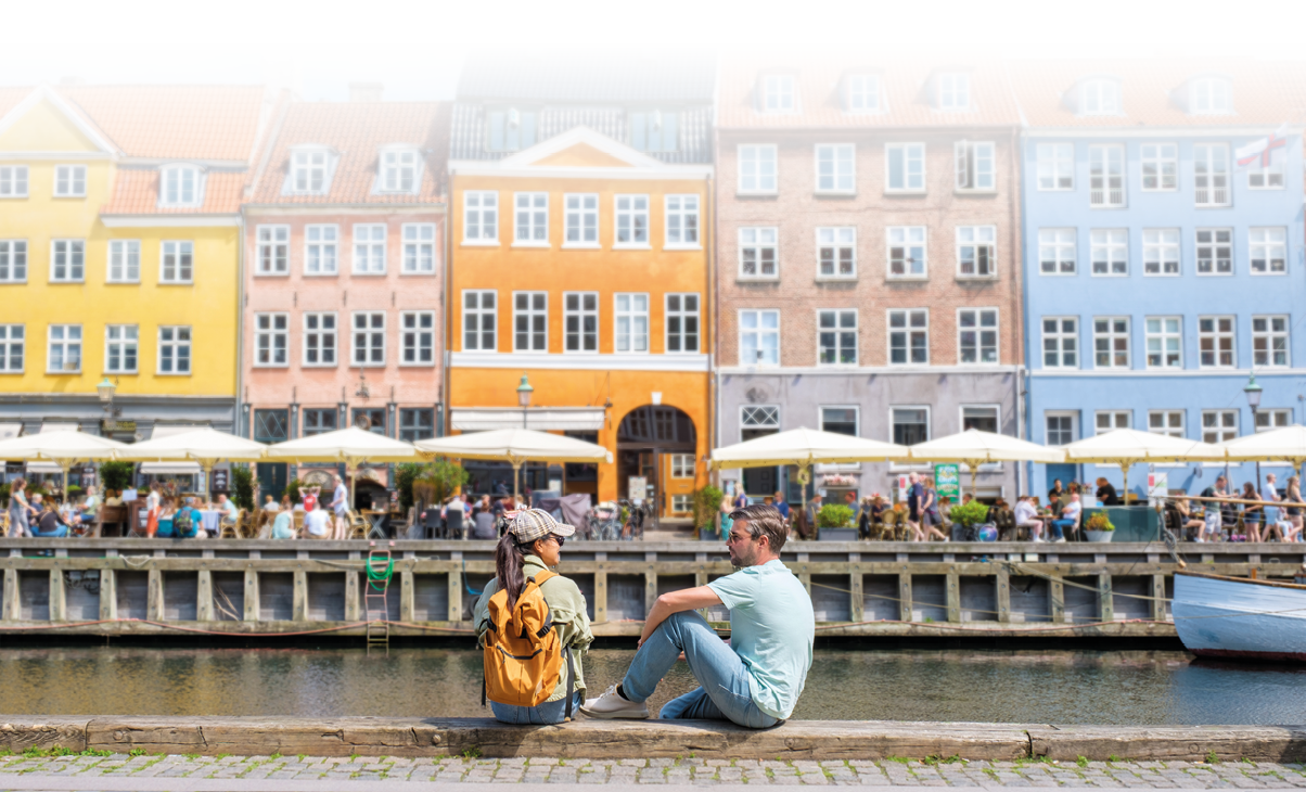 Zwei Personen sitzen am Ufer eines Kanals und unterhalten sich vor einer Reihe bunter, historischer H user mit Caf s und Menschen im Freien. Beschreibung von KI generiert