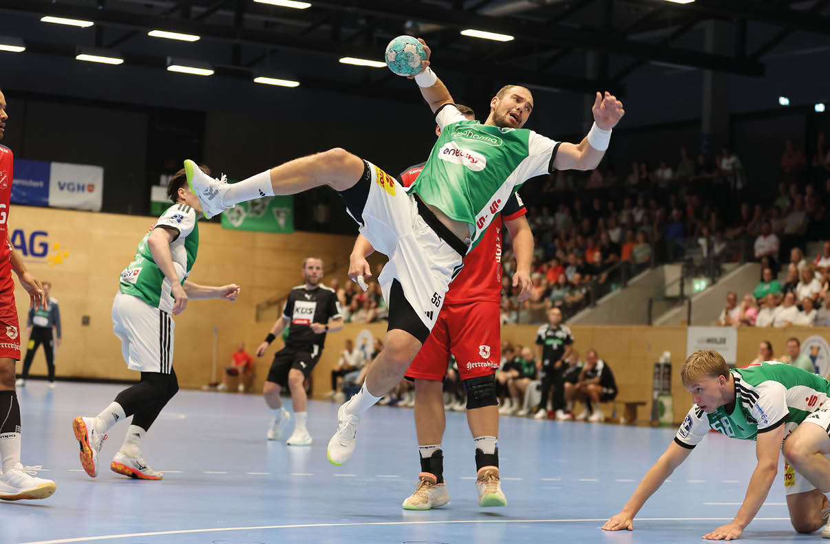 Mehrere Erwachsene spielen in einer Halle Handball, wobei eine Person in gr ner Sportkleidung in der Luft springt und den Ball h lt, w hrend andere Spieler in Bewegung sind. Beschreibung von KI generiert