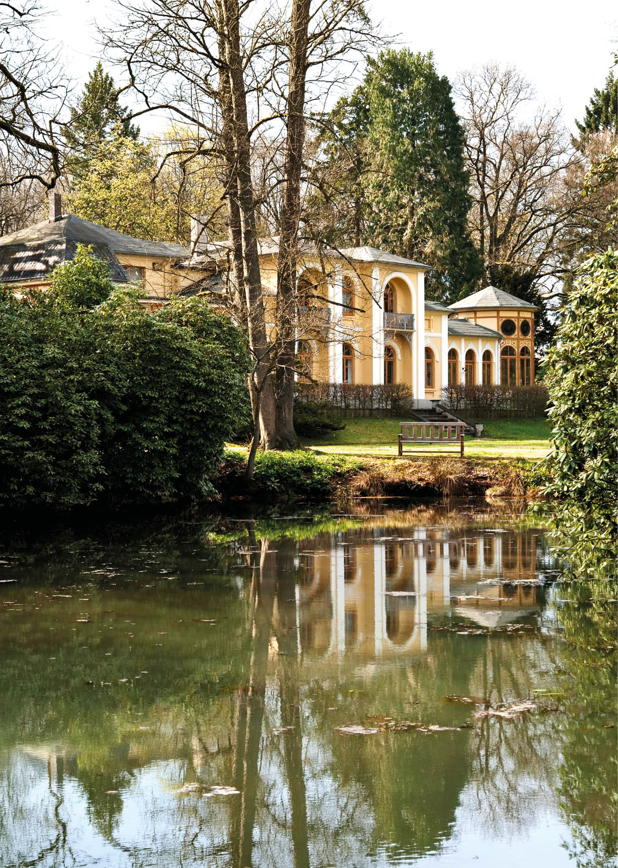 Ein historisches Geb ude mit S ulen und einem Pavillon steht hinter einem Teich, in dem sich das Bauwerk und die umliegenden B ume spiegeln. Beschreibung von KI generiert