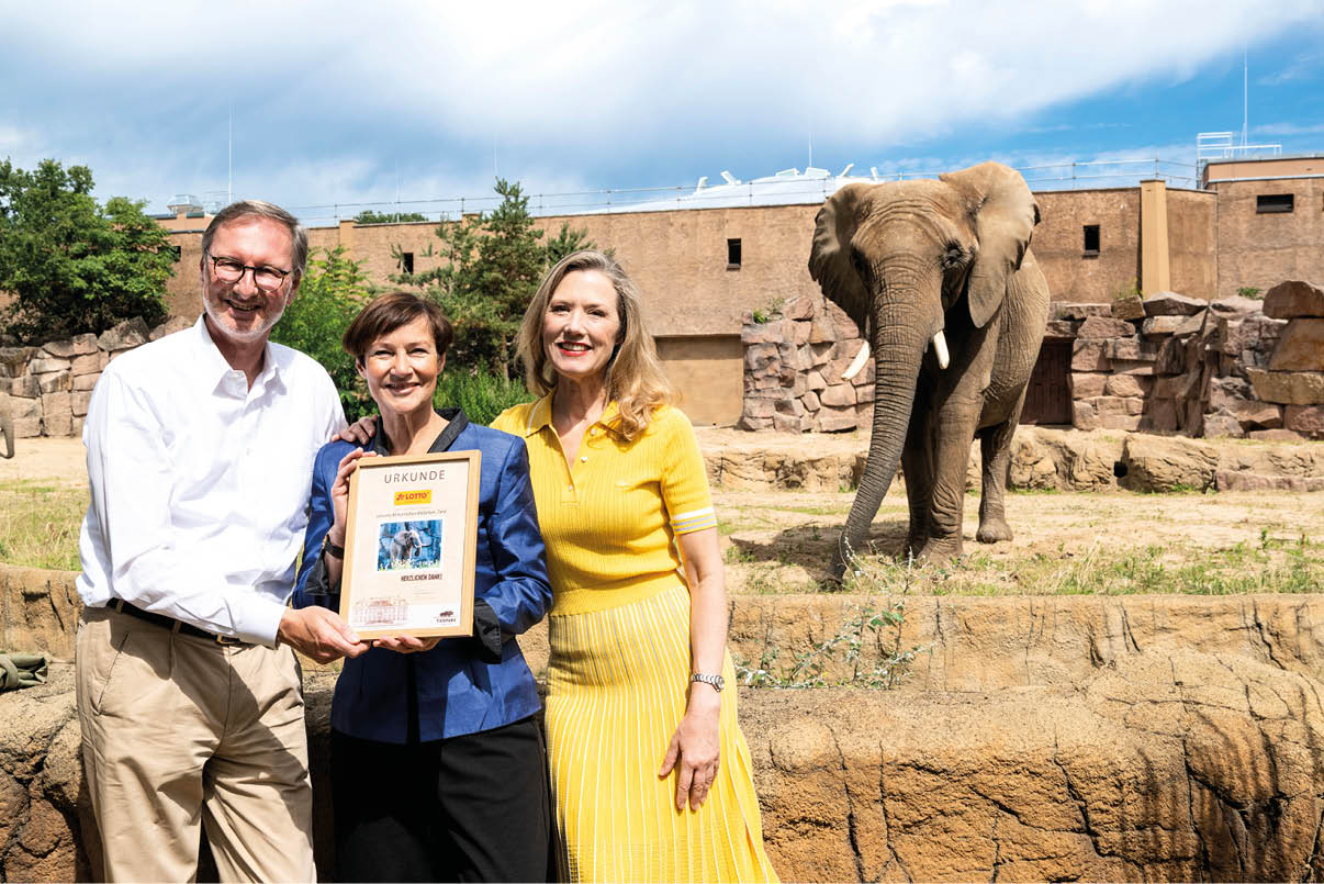 Drei Personen stehen l chelnd zusammen und halten eine Urkunde, w hrend im Hintergrund ein Elefant in einem Zoo-Gehge zu sehen ist. Beschreibung von KI generiert
