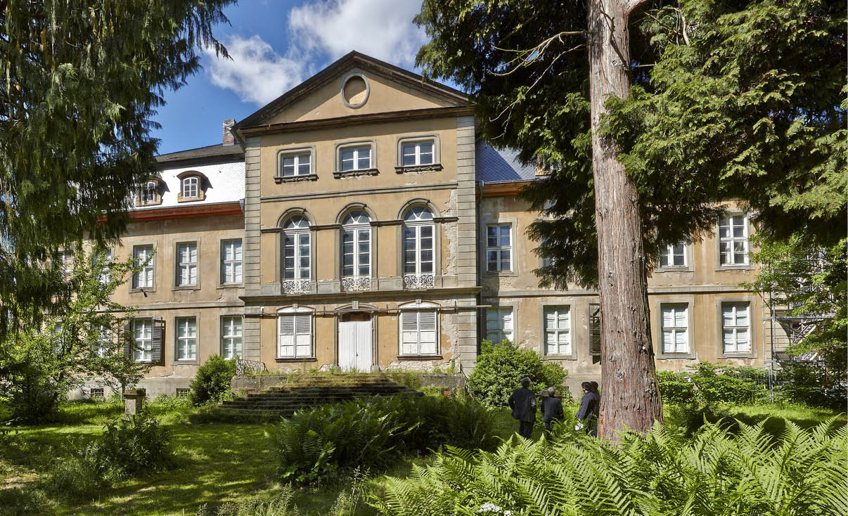 Ein gro es historisches Geb ude mit vielen Fenstern steht hinter einem gr nen Garten mit B umen und Farnen, w hrend zwei Personen nahe dem Geb ude stehen. Beschreibung von KI generiert