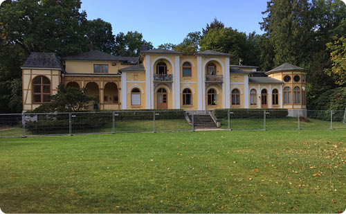 Ein gro es, historisch wirkendes Geb ude mit gelber Fassade und mehreren B gen steht hinter einem Zaun auf einer weitl ufigen gr nen Wiese, umgeben von hohen B umen. Beschreibung von KI generiert