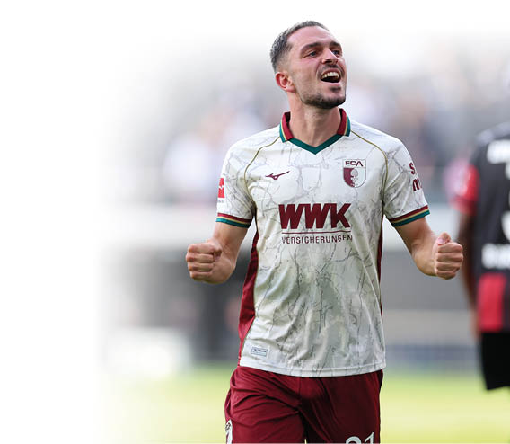 Ein jubelnder Spieler in wei -rotem Fu balltrikot steht auf dem Spielfeld, w hrend eine weitere Person in schwarzem Trikot mit der Nummer drei im Hintergrund zu sehen ist. Beschreibung von KI generiert