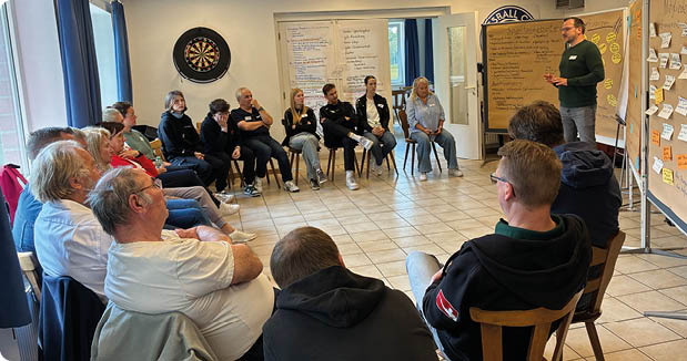 Eine Gruppe von Erwachsenen sitzt in einem Stuhlkreis in einem hellen Raum und h rt einer Person zu, die vor einer Pinnwand mit Notizen steht und spricht. Beschreibung von KI generiert