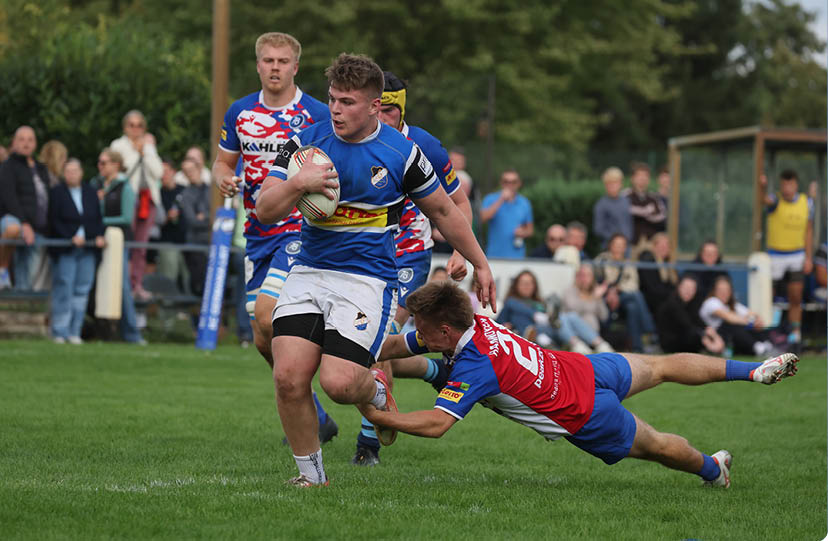 Mehrere Personen spielen auf einem Rasenplatz Rugby, wobei eine Person mit blauem Trikot den Ball h lt und eine andere Person im roten Trikot versucht, sie zu stoppen. Beschreibung von KI generiert