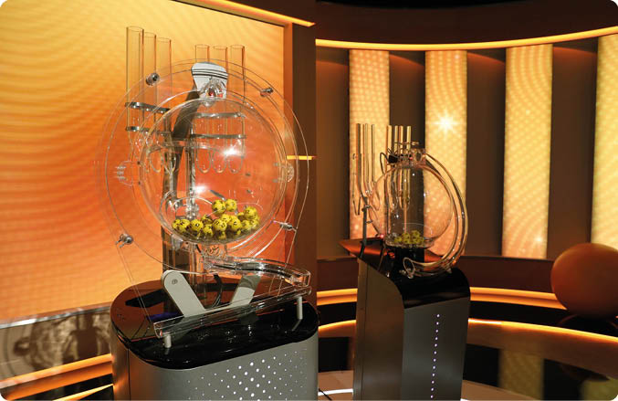 Zwei moderne Lotterie- oder Ziehungsmaschinen mit gelben Kugeln stehen in einem beleuchteten Studio mit warmem orangefarbenem Licht. Beschreibung von KI generiert