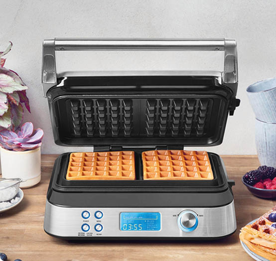 Ein moderner Waffeleisen mit zwei goldbraunen Waffeln steht ge ffnet auf einer Holzarbeitsplatte neben einer Pflanze, einer Tasse und einem Teller mit Beeren. Beschreibung von KI generiert