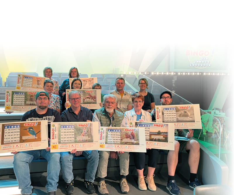 Eine Gruppe von Erwachsenen sitzt auf Trib nenstufen und h lt gro e Bingo-Karten in einer hell beleuchteten Halle mit blauem Hintergrund. Beschreibung von KI generiert