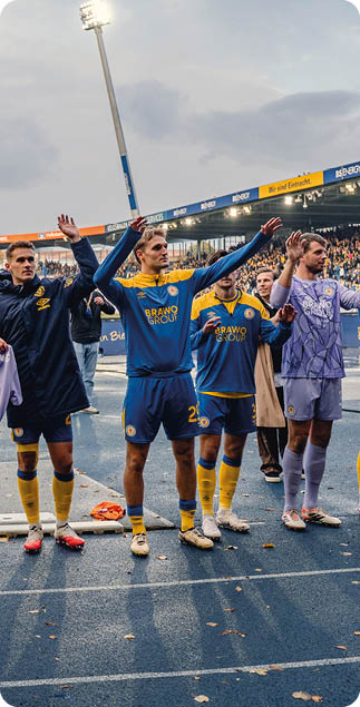 Eine Gruppe von Sportlern in gelben und blauen Trikots steht nebeneinander auf einer Laufbahn und hebt die Arme vor einem Stadion mit Zuschauern. Beschreibung von KI generiert
